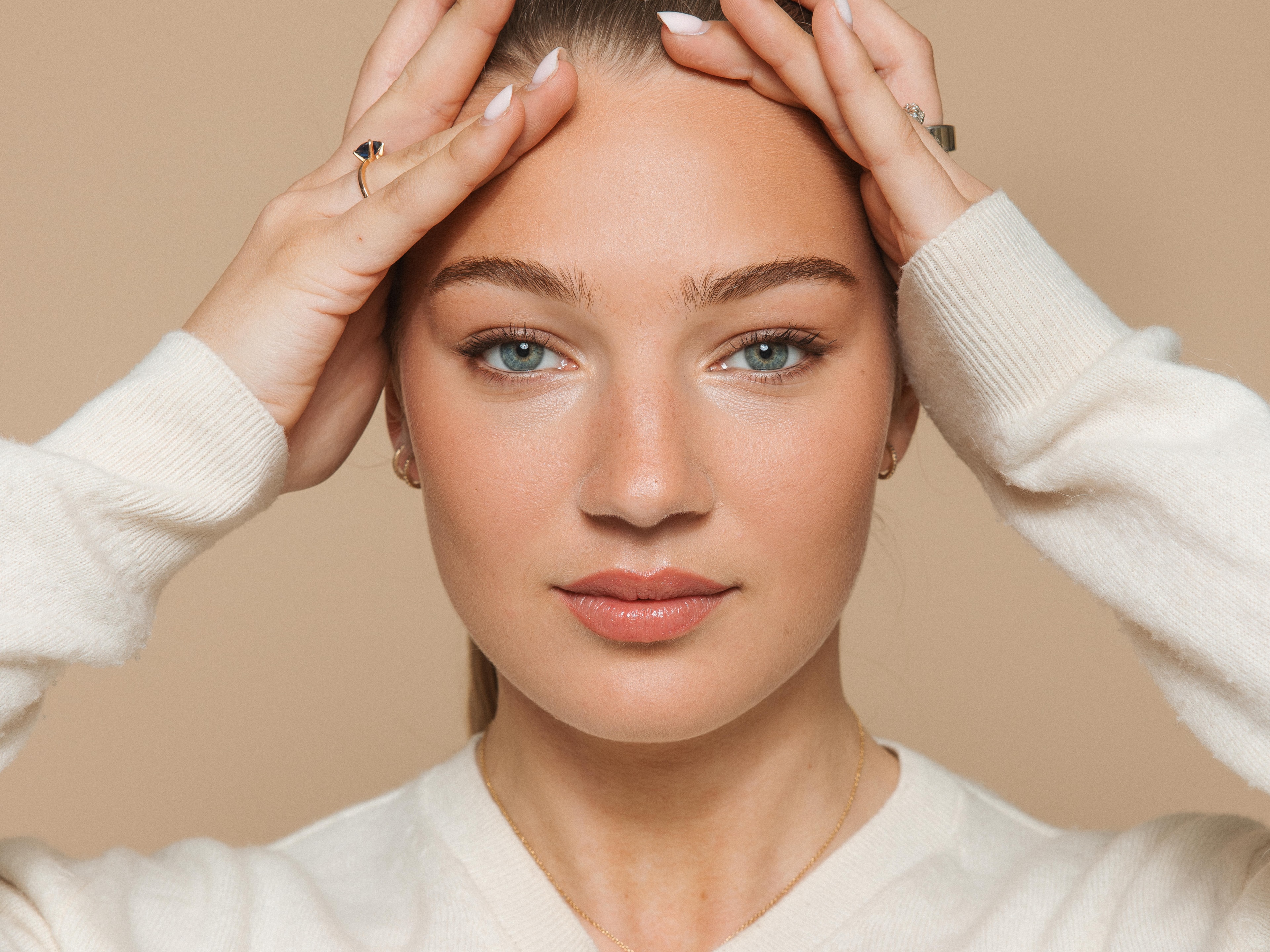 Soap Close-up portrait of a woman with natural makeup and blue eyes, holding her hands against her forehead while looking directly at the camera. She is wearing a cream-colored sweater and subtle jewelry against a beige background.